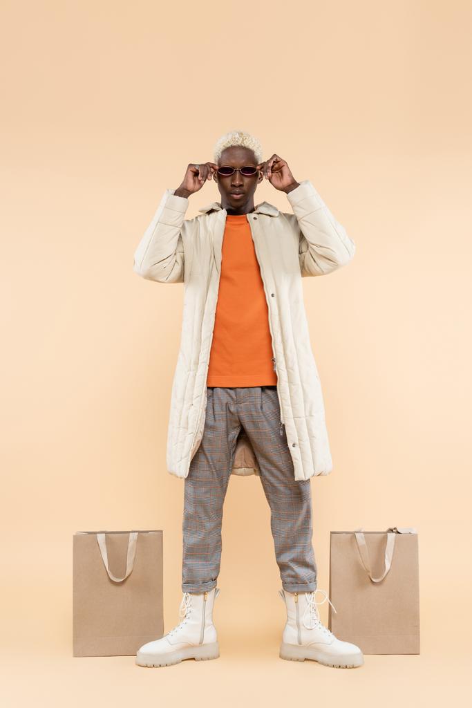 Modayı takip eden, şık montlu, bej rengi alışveriş torbalarının yanında güneş gözlüğü takan Afro-Amerikan bir adam. - Fotoğraf, Görsel