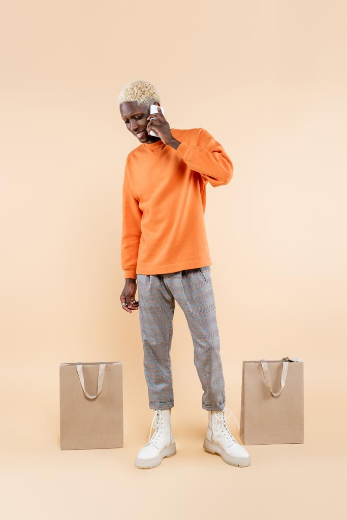 longitud completa de hombre afroamericano feliz en sudadera naranja hablando en el teléfono inteligente cerca de bolsas de compras en beige  - Foto, imagen