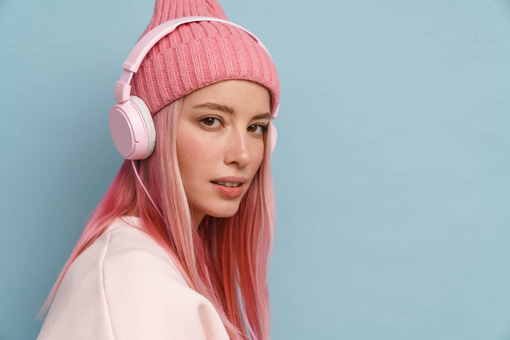 Mladá bílá žena nosí růžový klobouk poslech hudby s sluchátky izolované přes modrou stěnu - Fotografie, Obrázek