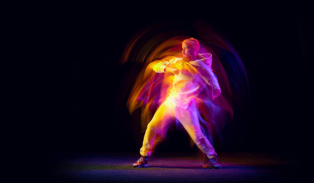 Baile en solitario. Joven hombre deportivo flexible bailando hip-hop o breakdance en traje blanco sobre fondo oscuro en luz mixta de neón amarillo. Belleza, deporte, juventud, acción, movimientos - Foto, imagen
