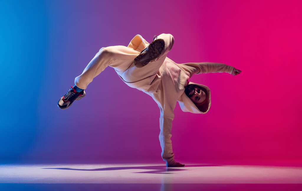 Photo studio de jeune homme sportif flexible dansant breakdance en tenue blanche sur fond rose bleu dégradé. Concept d'action, art, beauté, sport, jeunesse - Photo, image