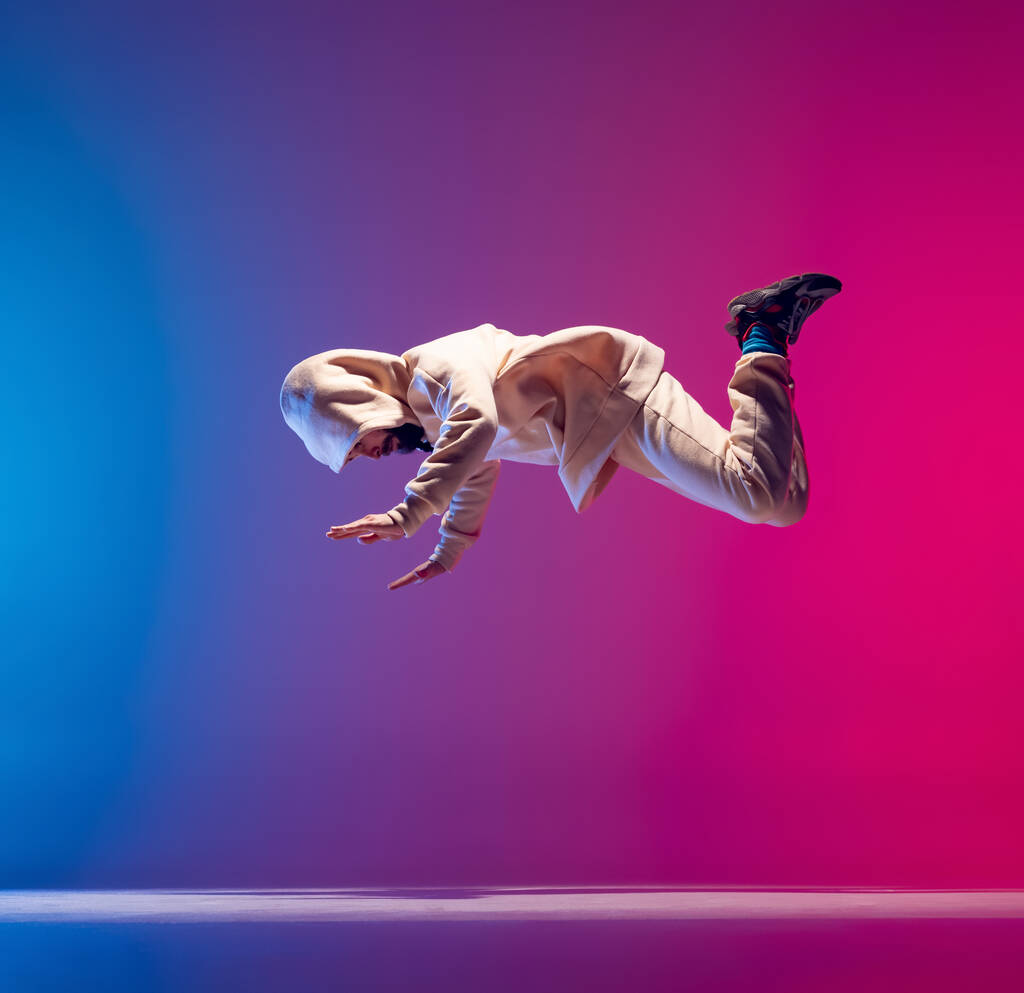 Freier Fall. Studioaufnahme eines jungen flexiblen, sportlichen Mannes, der Breakdance in weißem Outfit auf rosa-blauem Hintergrund tanzt. Handlungskonzept, Kunst, Schönheit, Sport, Jugend - Foto, Bild