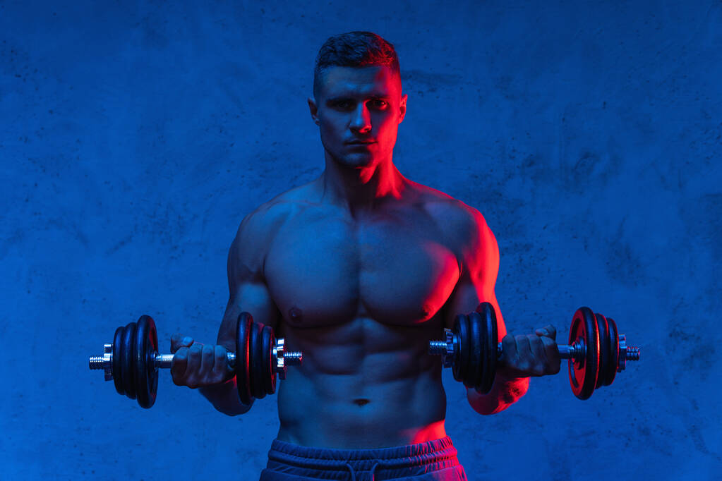 Young and muscular bodybuilder man exercising with dumbbells in colorful neon light - Photo, Image