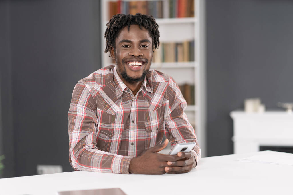 Hymyilevä komea Afrikkalainen Amerikkalainen mies käyttää älypuhelinta tarkistaa sosiaalisen median tai päivittää tietoja. Muotokuva nuoresta miehestä, jolla on älypuhelin pöydässä. Sosiaalisen median käsite - Valokuva, kuva
