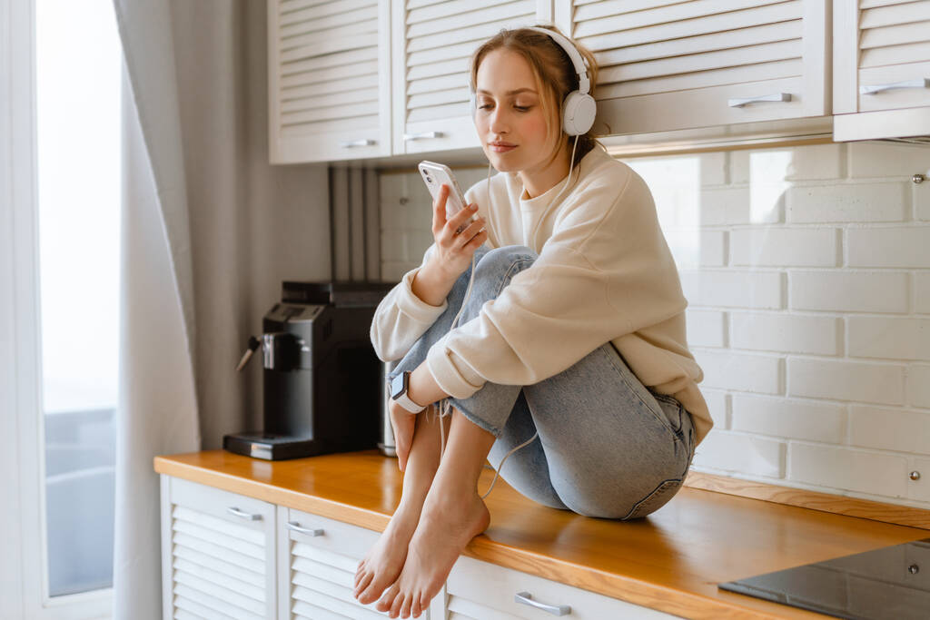 Fiatal nő zenét hallgat mobiltelefonnal és fülhallgatóval, miközben ül a konyhában - Fotó, kép