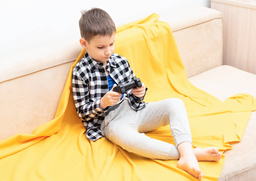 Der kleine Junge im karierten Hemd sitzt auf der Couch und spielt mit schwarzem Steuerknüppel in der Hand das Videospiel. Videospiele zu Hause spielen - Foto, Bild