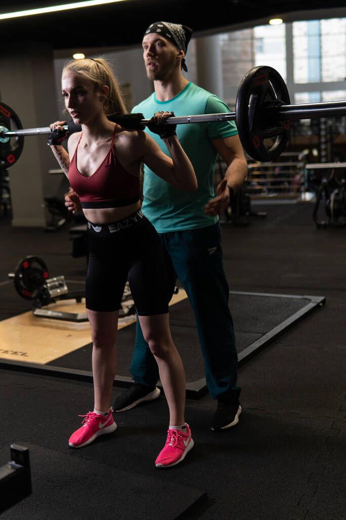 инструктор с клиенткой девушки, стоящей с штангой на плечах в красных красках в спортивном фитнес-клубе - Фото, изображение