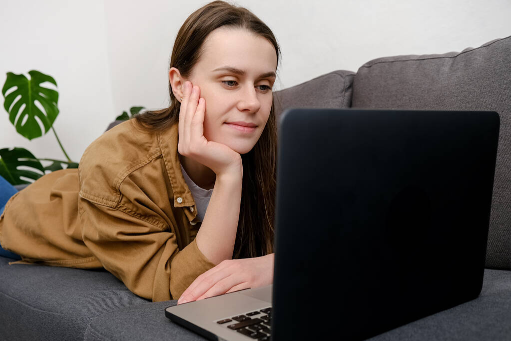 Schöne junge kaukasische Frau liegend auf der gemütlichen Couch zu Hause mit Laptop-Computer, Gerät Online-Suche, Fernstudium, Surfen im Internet beim E-Shopping entspannt auf bequemen grauen Sofa - Foto, Bild