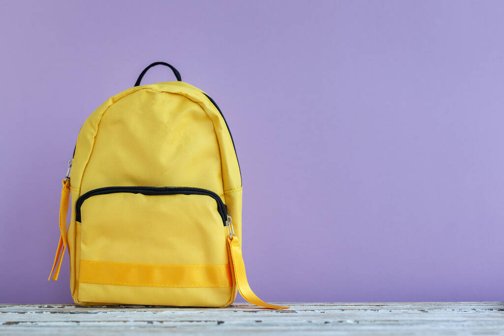 Ένα κίτρινο σακίδιο σε ένα άδειο μωβ φόντο με copyspace. Επιστροφή στο σχολείο έννοια. - Φωτογραφία, εικόνα