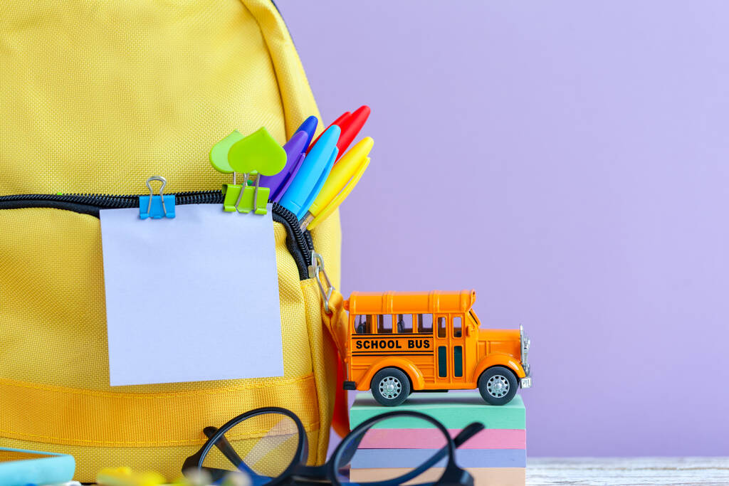 Concepto de vuelta a la escuela. Mochila escolar amarilla completa y escuela de autobús de juguete con papelería en la mesa sobre fondo púrpura. - Foto, imagen