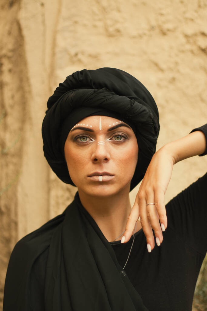 Retrato de una joven hermosa mujer con maquillaje y adorno de arte de cara blanca en estilo tribal o de Oriente Medio vestida con un hiyab negro. Imagen étnica Primer plano sobre el fondo de una pared arenosa. - Foto, imagen