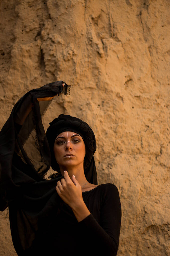 Portrait of a young beautiful woman with make-up and white face art ornament in tribal or middle eastern style dressed in a black hijab, dancing. Ethnic image on the background of the sandy wall - Foto, Imagem