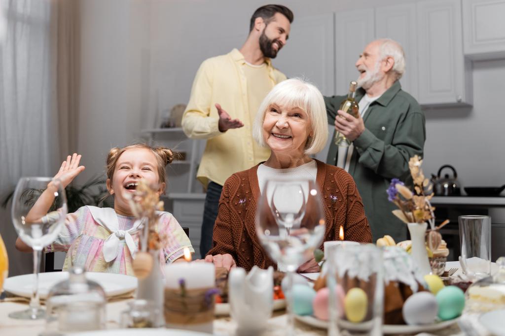 podekscytowana dziewczyna macha ręką w pobliżu uśmiechniętej babci podczas uroczystej kolacji w domu - Zdjęcie, obraz