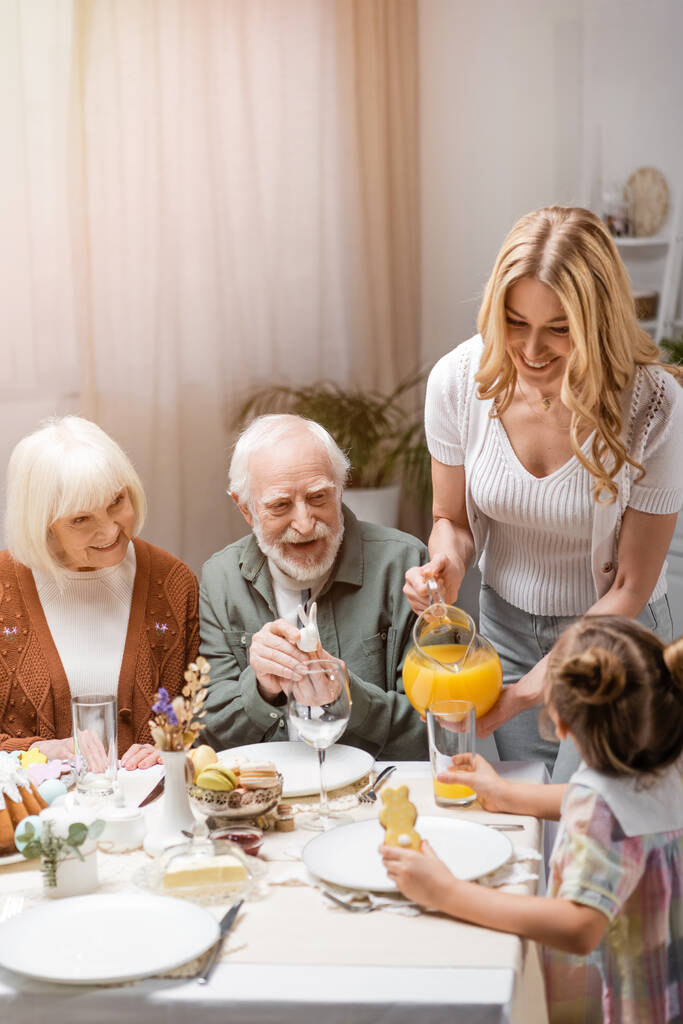 Lächelnde Frau schenkt Orangensaft ein, während Familie zu Hause ein Osteressen einnimmt - Foto, Bild