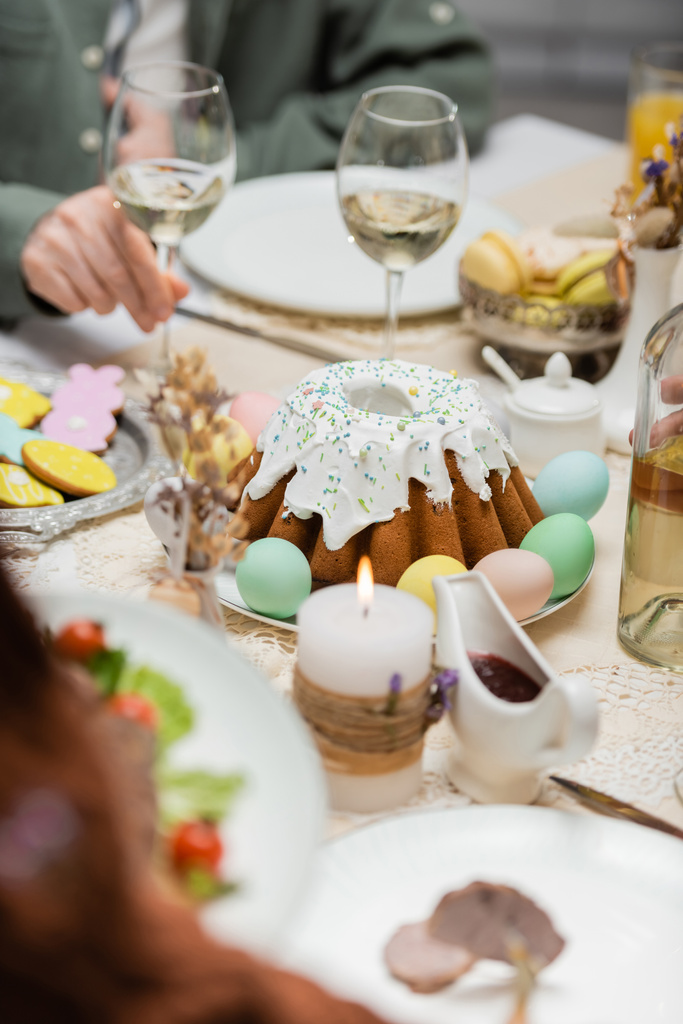 Teilansicht des Menschen bei leckerem Osterkuchen, bemalten Eiern und Weingläsern auf festlichem Tisch - Foto, Bild