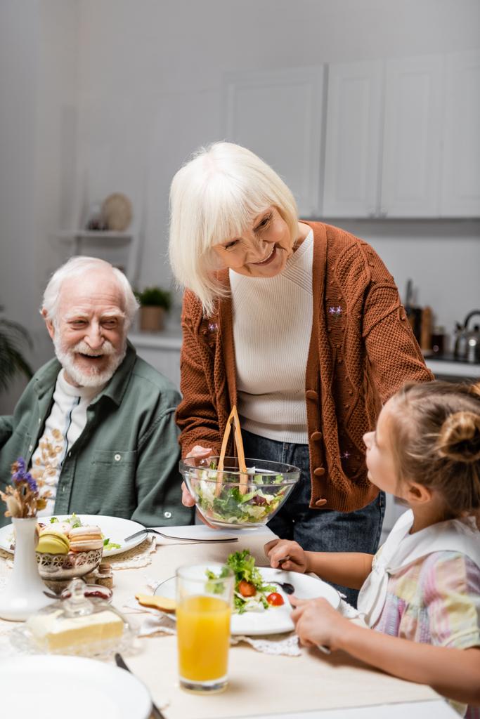 donna anziana sorridente che tiene l'insalata vegetale vicino a nipote durante cena pasquale - Foto, immagini