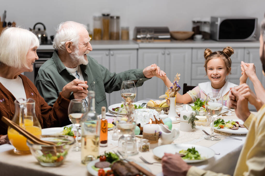 イースターディナーでテーブルに座りながら手をつなぐ陽気な家族 - 写真・画像