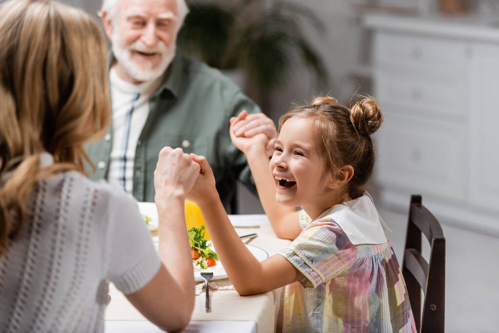 excitada chica cogida de la mano con borrosa mamá y el abuelo durante la cena de Pascua - Foto, Imagen