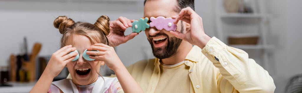 γελαστοί πατέρας και κόρη που καλύπτουν τα μάτια με πασχαλινά μπισκότα και ζωγραφισμένα αυγά ενώ διασκεδάζουν στο σπίτι, πανό - Φωτογραφία, εικόνα