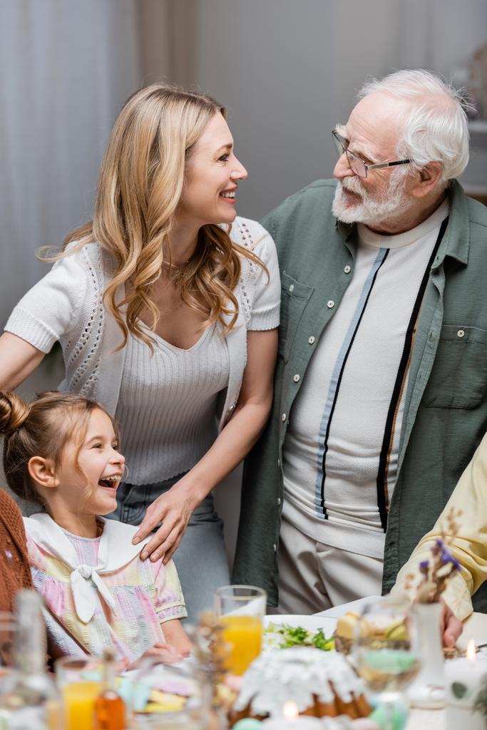 happy woman talking to senior dad near laughing daughter during easter dinner - Photo, Image