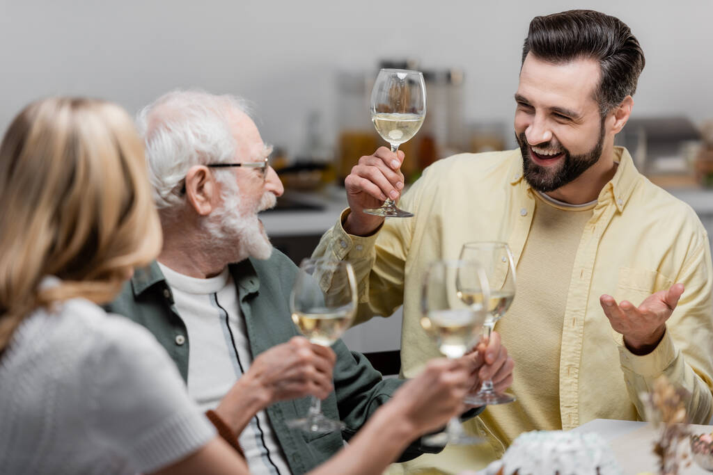 Senior hält ein Glas Wein in der Hand, während er mit erwachsenen Söhnen und Töchtern Ostern feiert  - Foto, Bild