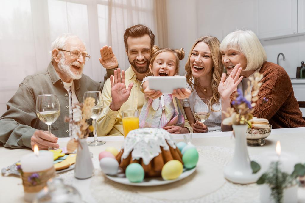 веселая семья машет руками во время видеозвонка на смартфон рядом со столом с пасхальным ужином - Фото, изображение