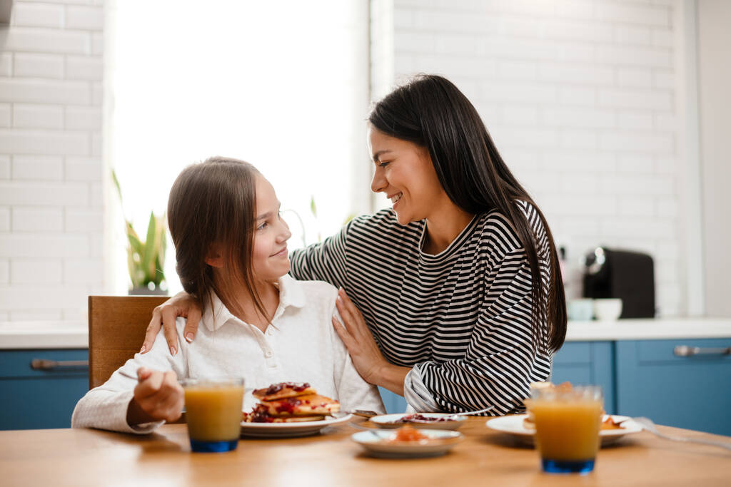 Cheerful beautiful mother and daughter hugging while having breakfast in cozy kitchen - Photo, Image