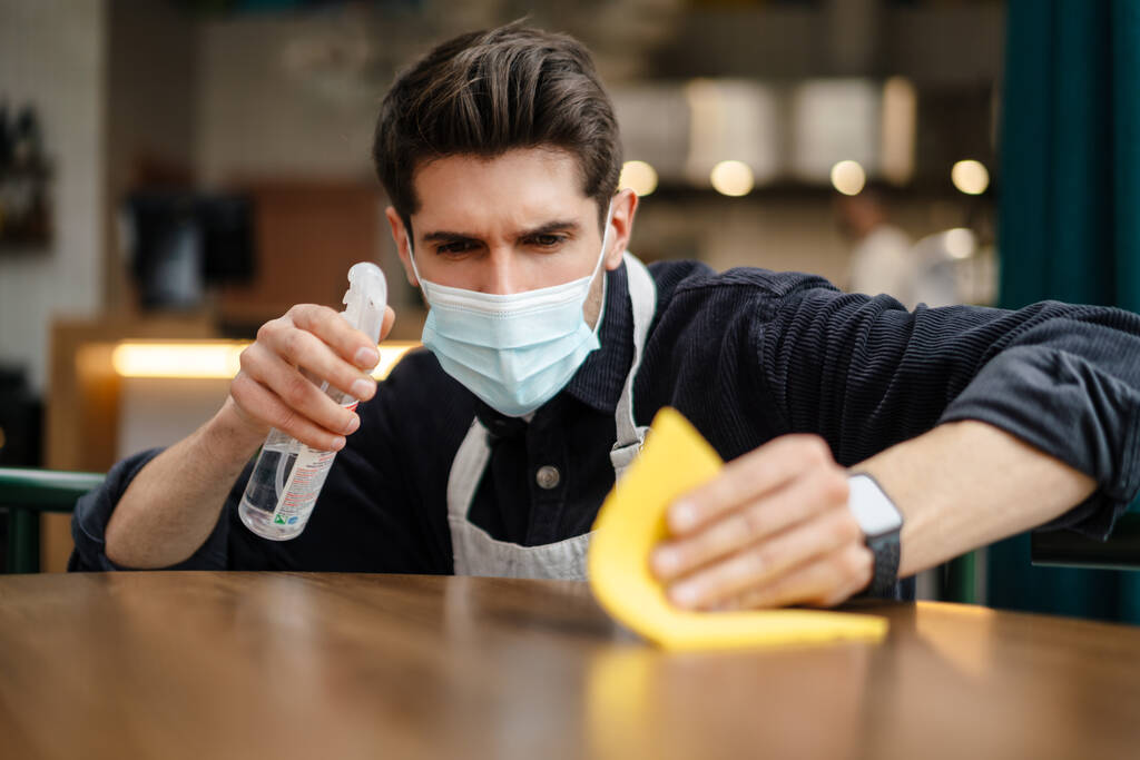 Man ober met masker desinfecterende tafel in het cafe binnen, close-up - Foto, afbeelding