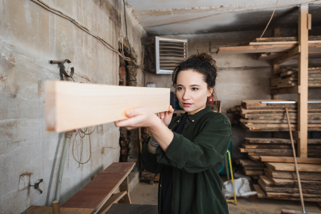 Молодой плотник смотрит на деревянную доску в мастерской  - Фото, изображение