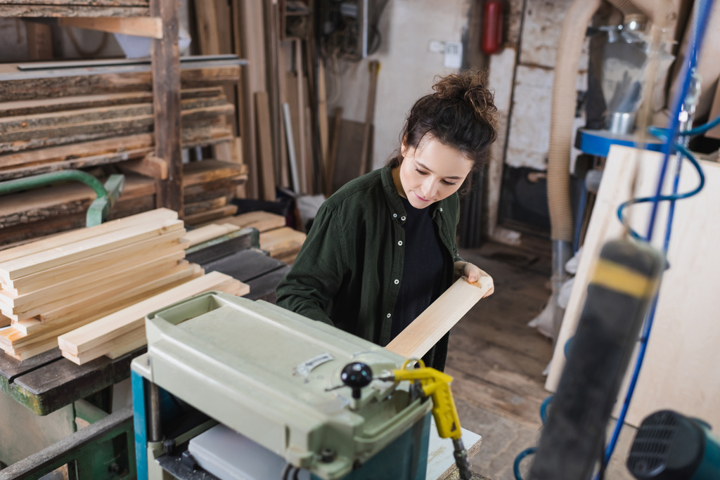 Brünetten Tischler hält Holzbrett in der Nähe Dicke Hobelmaschine und Bohlen in Werkstatt  - Foto, Bild