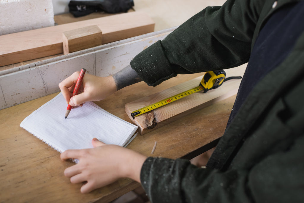 Обрізаний вид тесляра, що пише на блокноті біля дерев'яної дошки та вимірювання стрічки в майстерні
  - Фото, зображення