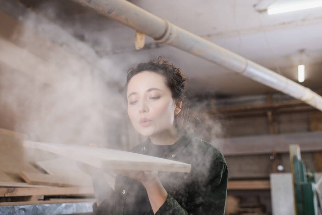Плотник выдувает опилки из деревянной доски в мастерской  - Фото, изображение