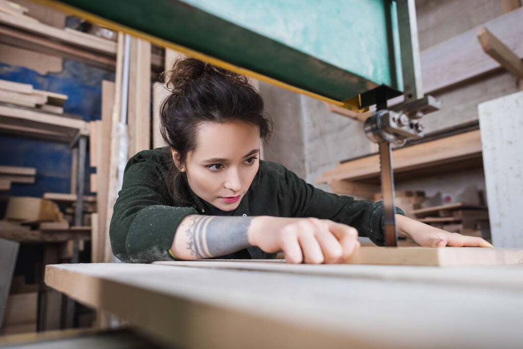 Morena carpinteiro segurando placa de madeira borrada e trabalhando em serra de banda na oficina  - Foto, Imagem