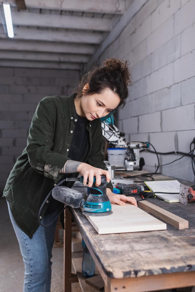 Brunette charpentier ponçage planche de bois près des outils en atelier  - Photo, image