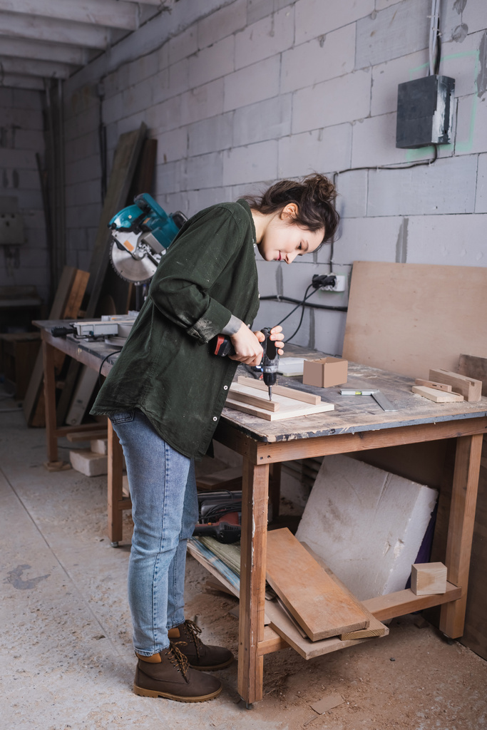 Вид сбоку на молодого дизайнера мебели, работающего с электрической дрелью и деревянной доской в мастерской  - Фото, изображение