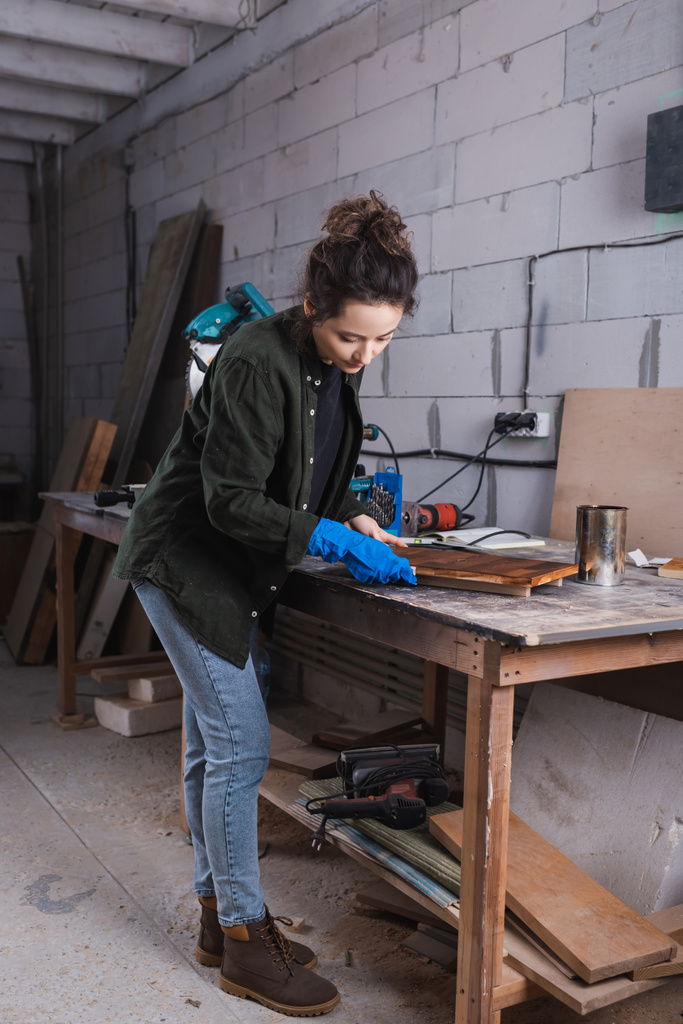 Jeune menuisier en gant de caoutchouc appliquant une tache de bois sur la planche près du bocal et de la scie à onglets en atelier  - Photo, image