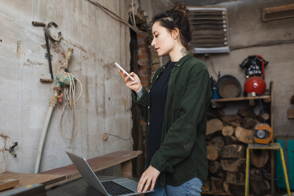 Вид сбоку на татуированного деревообработчика, использующего мобильный телефон рядом с ноутбуком в мастерской  - Фото, изображение
