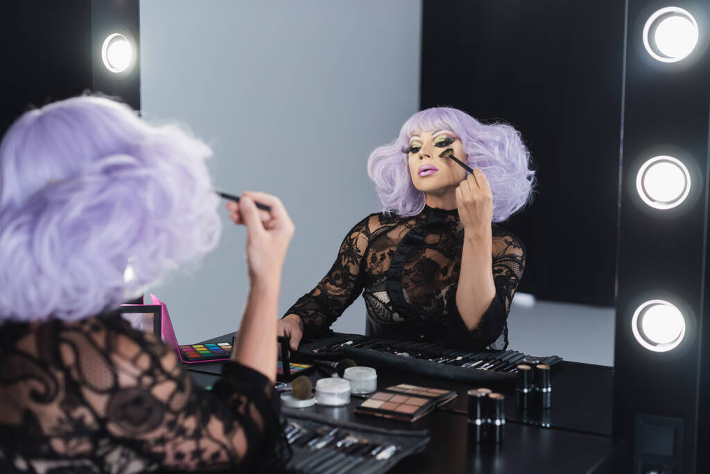 τρανσέξουαλ πρόσωπο σε βιολετί περούκα εφαρμογή μακιγιάζ, ενώ κοιτάζοντας στον καθρέφτη - Φωτογραφία, εικόνα