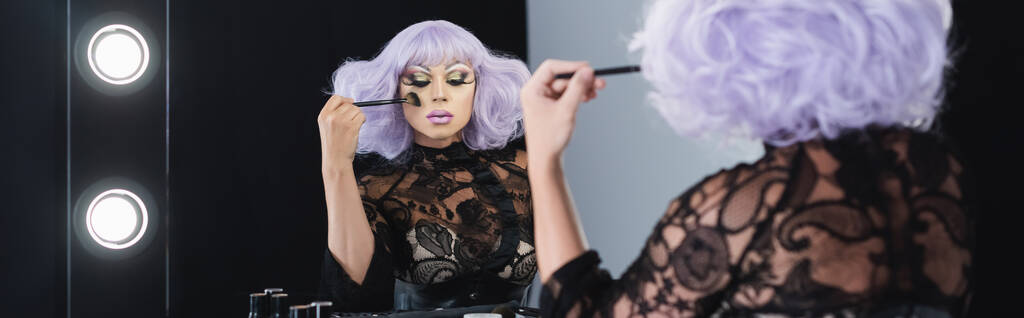 hombre excéntrico en peluca púrpura aplicando polvo facial cerca del espejo, pancarta - Foto, Imagen