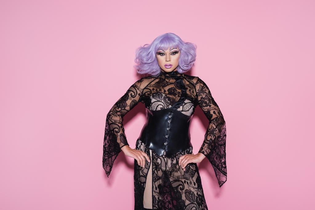 excéntrica drag queen en peluca púrpura y vestido de encaje negro mirando a la cámara en rosa - Foto, Imagen