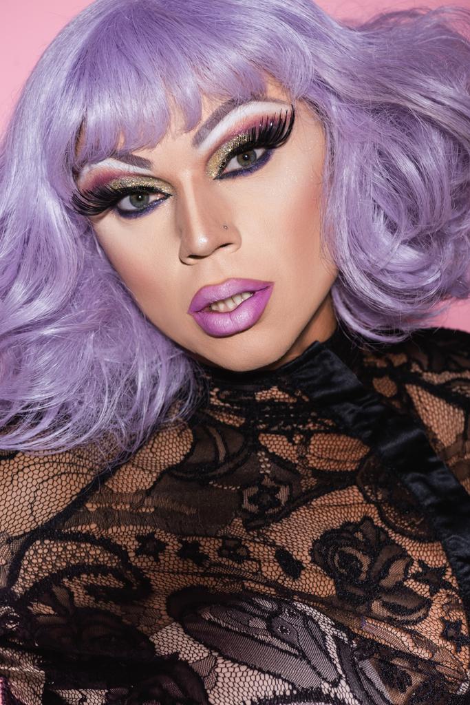 excéntrico hombre de peluca púrpura y maquillaje brillante mirando a la cámara aislada en rosa - Foto, imagen