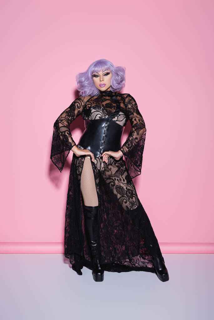full length άποψη του glamour drag queen σε μωβ περούκα και φόρεμα δαντέλα στέκεται με τα χέρια στους γοφούς σε ροζ - Φωτογραφία, εικόνα