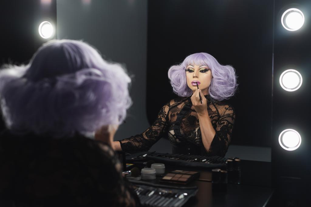 гламурная трансвестит в фиолетовом парике нанося контур губ косметическим карандашом - Фото, изображение