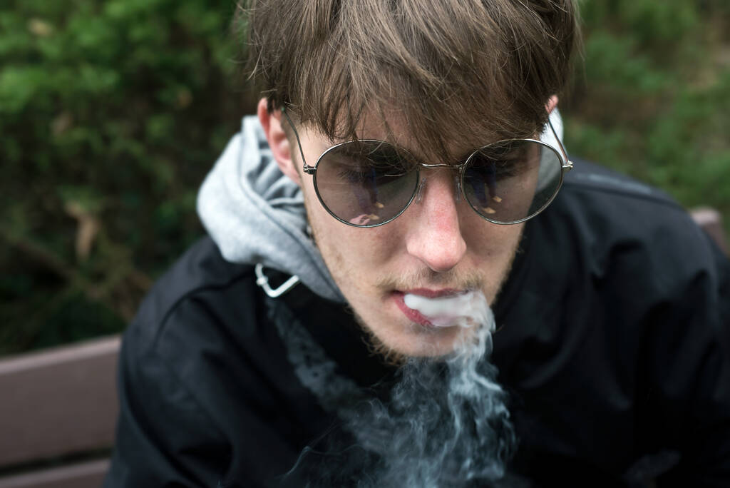 Portret van een jonge man met een zonnebril die een sigaret rookt op straat - Foto, afbeelding