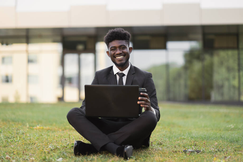 Νεαρός Αφρικανός επιχειρηματίας που φοράει μαύρο κοστούμι κάθεται στο γρασίδι με laptop και παίρνει τον καφέ. - Φωτογραφία, εικόνα