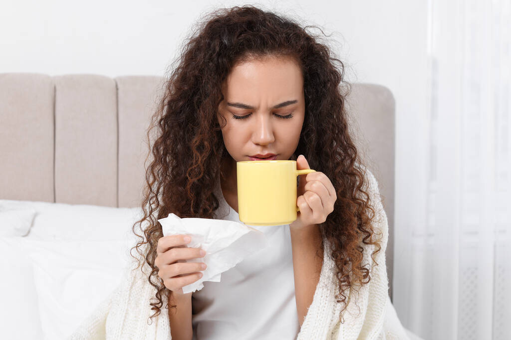 Femme afro-américaine malade avec des tissus et une tasse de boisson au lit à la maison - Photo, image