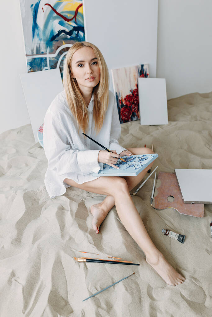 Holka sedí na písku ve foto studiu. Mladý umělec maluje obraz ve studiu. - Fotografie, Obrázek