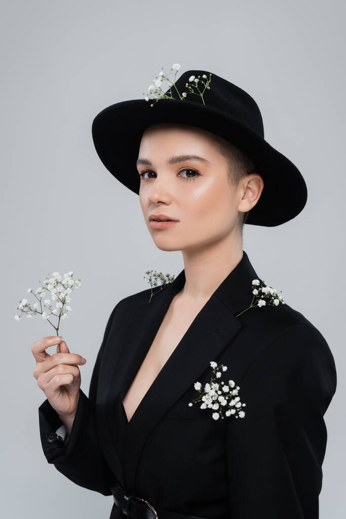 mujer con pequeñas flores blancas en blazer negro y sombrero fedora mirando a la cámara aislada en gris - Foto, imagen