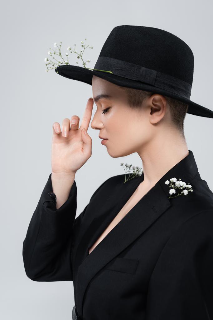 side view of woman with closed eyes pointing at black brim hat with gypsophila flowers isolated on grey - Photo, Image