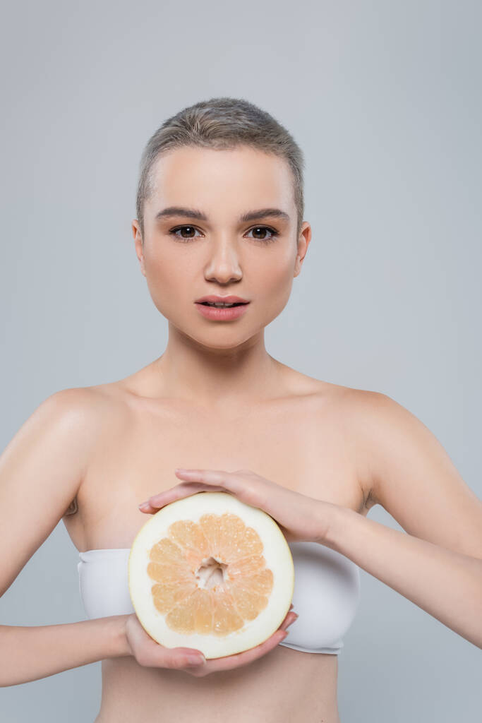 Charmante Frau mit perfekter Haut und nackten Schultern, die reife Grapefruit isoliert auf grau hält - Foto, Bild
